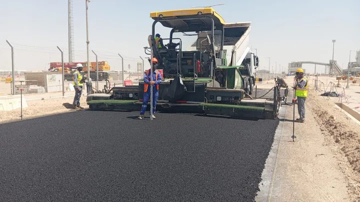 فنيشر أسفلت وفريق عمل يفرشان طبقة سوداء على الطريق.